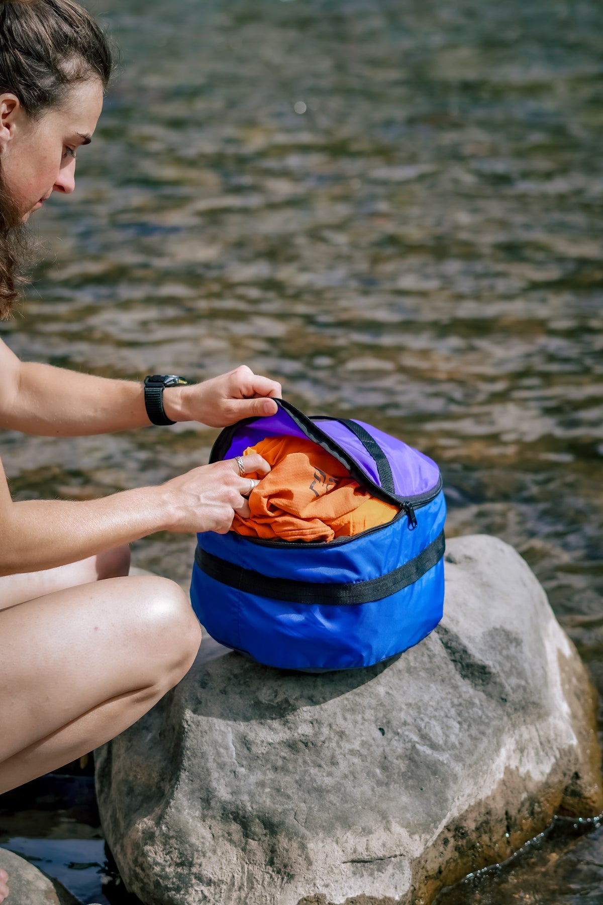 Person unzipping a Ruby Moons packing round on a river rock, with an orange garment packed inside
