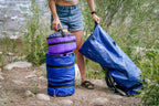 Person by a river holding a blue dry bag open, beside three stacked Ruby Moons packing rounds on rocky ground