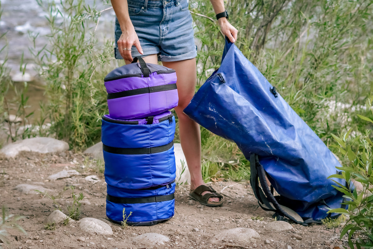 Person by a river holding a blue dry bag open, beside three stacked Ruby Moons packing rounds on rocky ground