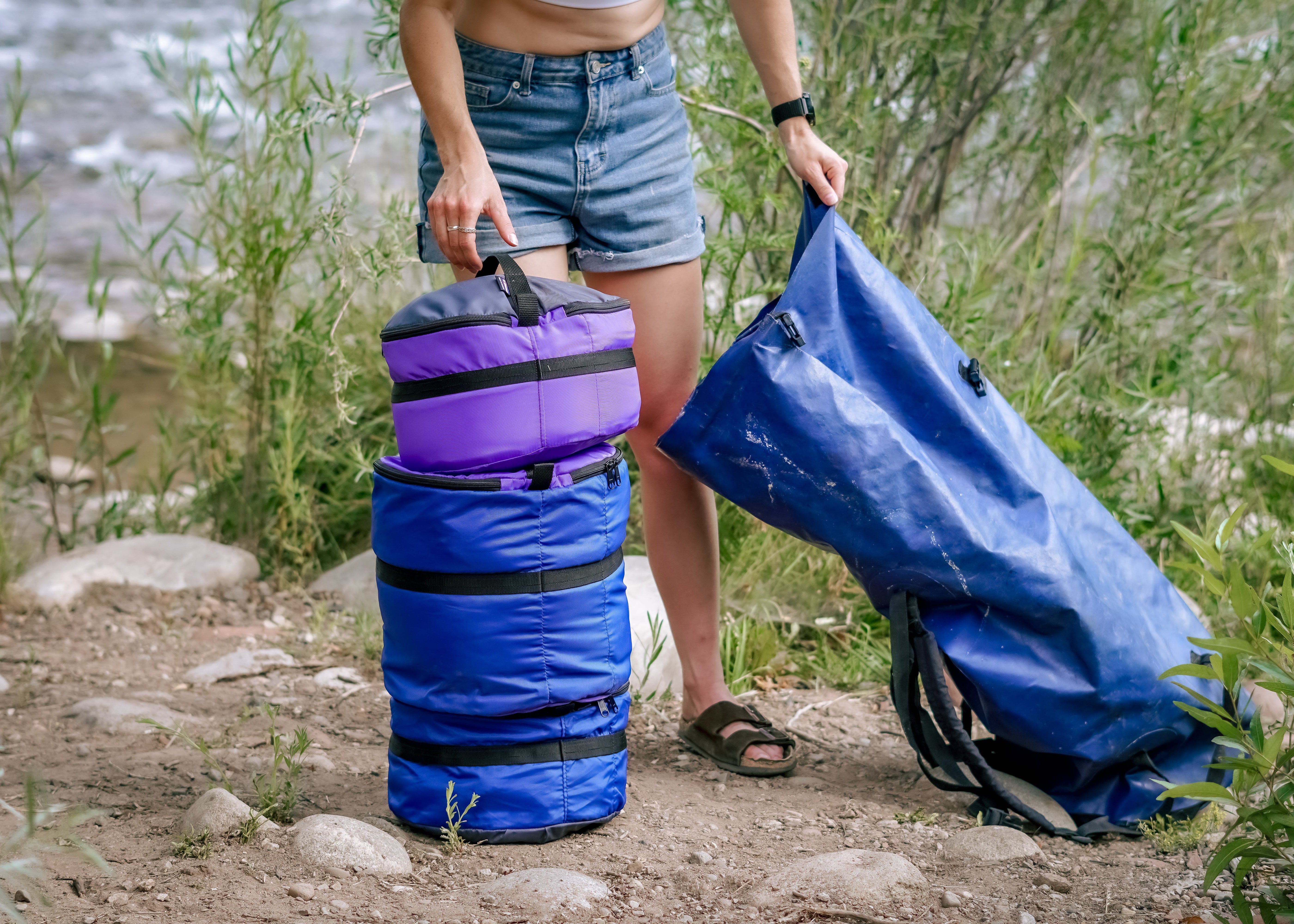 A person outdoors by a river or stream stands next to two waterproof dry bags: a purple and blue stack on the left, and a large blue bag being held open on the right.