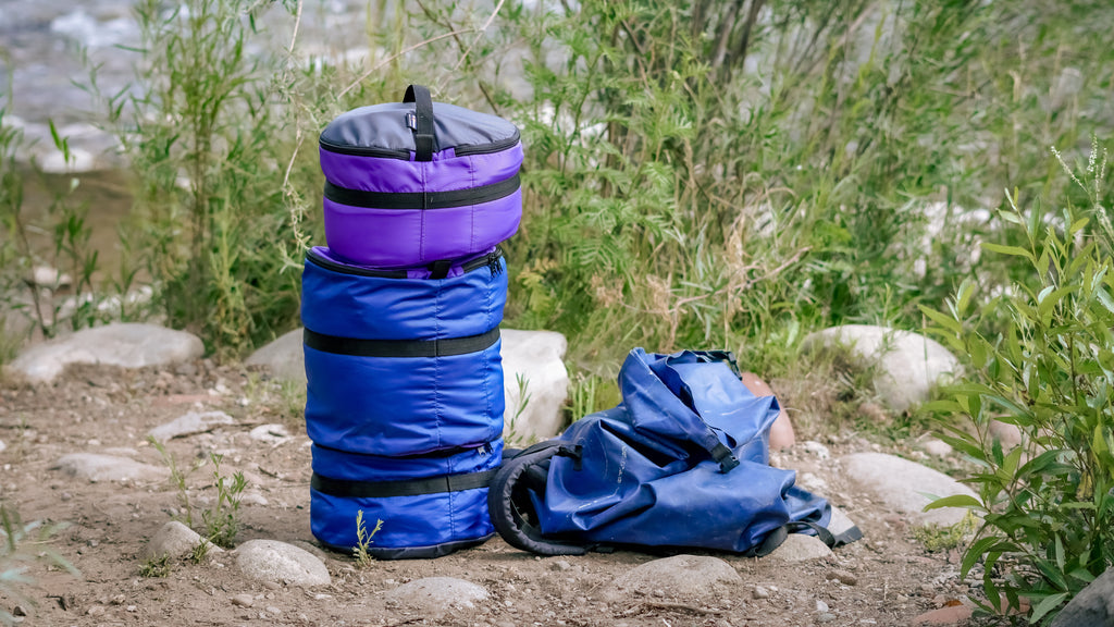 Three Ruby Moons packing rounds stacked in purple and blue, beside a collapsed blue dry bag, on riverside rocks and dirt