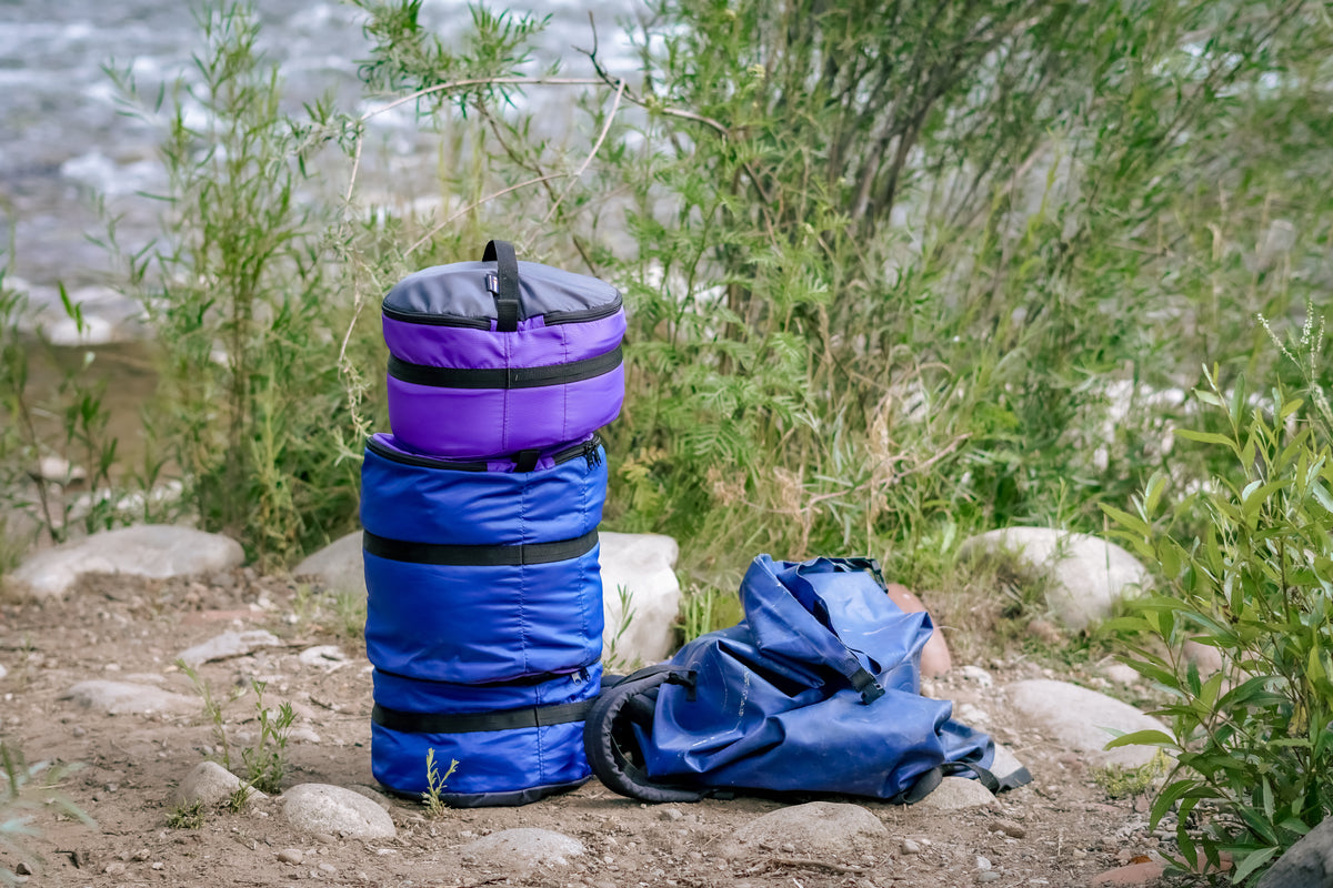 Three Ruby Moons packing rounds stacked in purple and blue, beside a collapsed blue dry bag, on riverside rocks and dirt