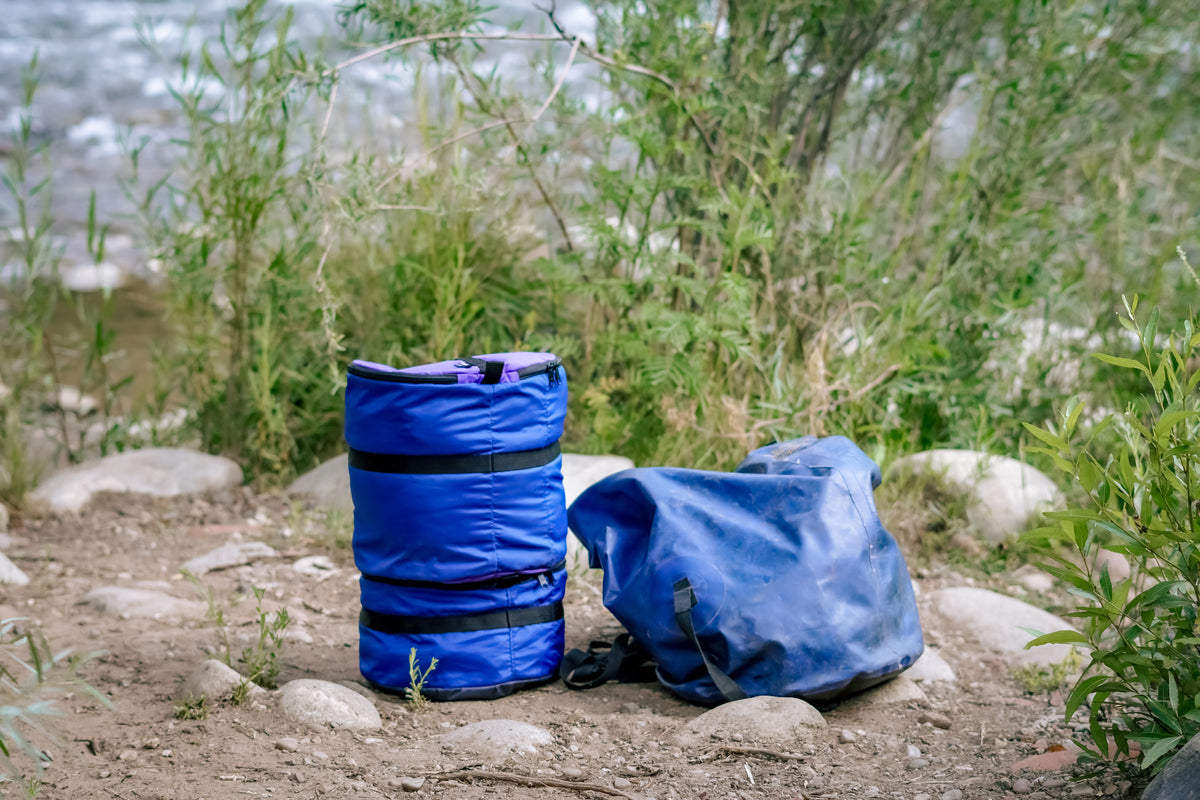 Two Ruby Moons packing rounds stacked in purple and blue, beside a collapsed blue dry bag, on riverside rocks and dirt