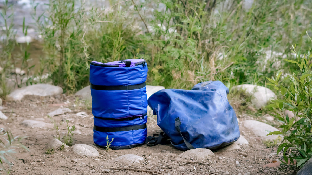 Two Ruby Moons packing rounds stacked in purple and blue, beside a collapsed blue dry bag, on riverside rocks and dirt