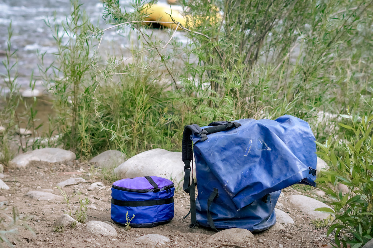 One Ruby Moons packing round stacked in blue, beside a collapsed blue dry bag, on riverside rocks and dirt