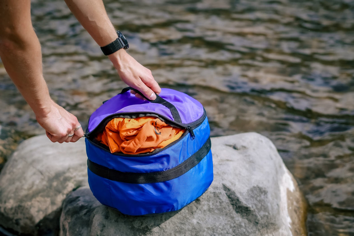 Person unzipping a Ruby Moons packing round on a river rock, with an orange garment packed inside